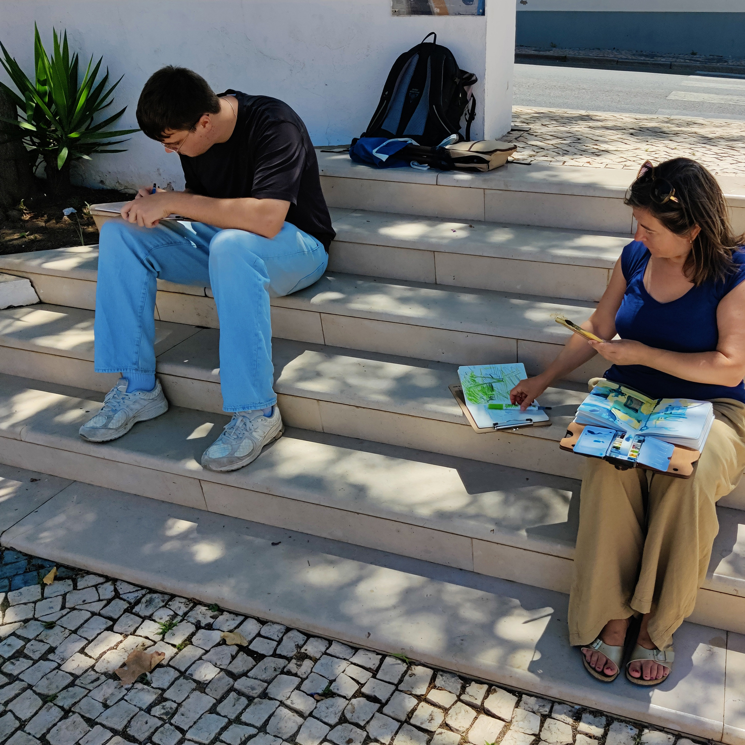 Sketching Moments in Alcoutim – Outdoor Watercolor Practice Two people watercolor painting in sketchbooks while sitting on shady outdoor steps in Alcoutim, Portugal.