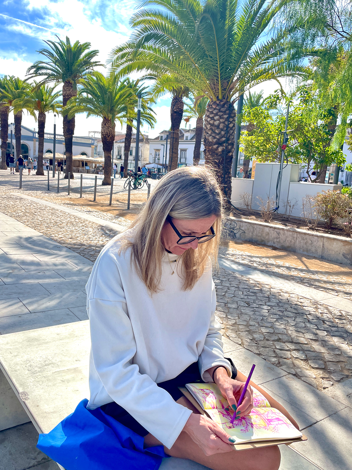 Woman sitting in front of the Mercado de Tavira, sketching with the market entrance and palm trees visible in the background.