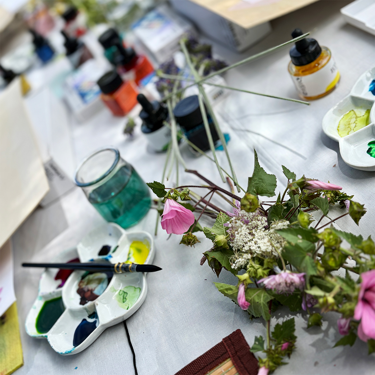 Mixed-media painting materials prepared outdoors with fluid inks, watercolour palettes, brushes and freshly picked wildflowers.